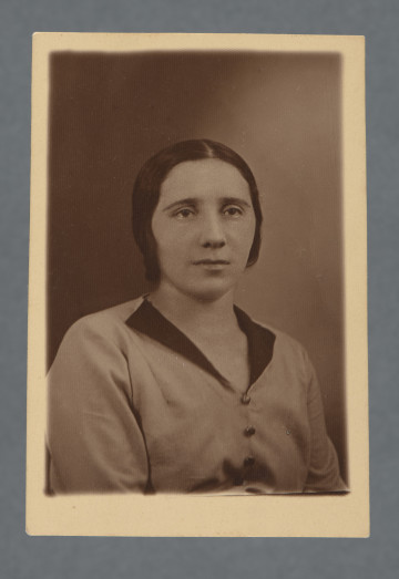 MPOLIN-A48.1.28 Monochromatyczna fotografia studyjna kobiety, popiersie, ubranej w średniojasną suknię (?) z ciemnym kołnierzem i wyraźnymi guzikami, dekolt trójkątny. Włosy z przedziałkiem, płasko przy głowie opadające za uszy, półuśmiech.Krawędzie proste, z marginesem - przy dolnej krawędzi większym niż przy innych.