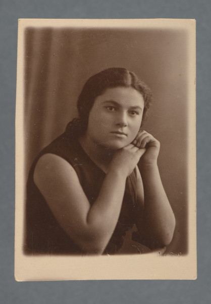 Fotografia gabinetowa, popiersie, kobiety z brodą podpartą na dłoni, w ciemnej bluzce bez rękawów, włosami upiętymi z tyłu, lokowanymi, z przedziałkiem. Twarz ujęta w trzech czwartych, nie patrzy w obiektyw, ale za niego. Na tle ciemnej kotary (?).Fotografia z białym wąskim marginesem, większym u dołu, wzdłuż prostych krawędzi.