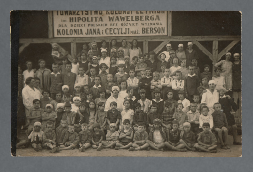 Grupa wielu osób w co najmniej siedmiu rzędach, zgromadzonych przed drewnianą konstrukcją z wielkim napisem: ,,TOWARZYSTWO KOLONJI LETNICH | im. HIPOLITA WAWELBERGA | DLA DZIECI POLSKICH BEZ RÓŻNICY WYZNANIA | KOLONIA JANA I CECYLJI MAŁŻ. BERSON''. Fotografia wychowanków i kadry (zapewne to kadra ma w większości białe chusty na głowach) kolonii letnich dla dzieci, które organizowano rokrocznie w Ciechocinku oraz - jak zapewne w tym wypadku - w Lesznie pod Warszawą (co wiemy z np. jednoniówki ,,Dzieci o wakacjach. Jednodniówka wydana staraniem Tow. Kolonji Letnich im. H. Wawelberga dla Dzieci Polskich bez Różnicy Wyznania, Warszawa, wrzesień 1929, zob. s. 2; egz. w zbiorach BN, sygn. DŻS XVIIA 9a).Krawędzie proste, w prawych rogach widoczne przebarwienia.