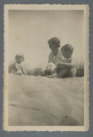 Fotografia przedstawia dwoje dzieci i kobietę na piasku, w tle trawy - być może nieopodal wody (rzeki, jeziora, morza)? Na rewersie dedykacja po niemiecku, z datą 1936, od Elli Busilmann (?).Najprawdopodobniej jedna z pamiątek Hilela Poznera, emigrującego w 1935 r. do Palestyny.Krawędzie ząbkowane.