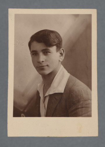 Fotografia monochromatyczna, w sepii, przedstawia uśmiechniętego młodego mężczyznę, tego samego i zapewne w tej samej marynarce i koszuli co na zdjęciu wcześniejszym, w klapie marynarki nie ma jednak, w odróżnieniu od tamtego, przypinki.Kołnierzyk - duży - białej koszuli wyłożony na ciemną (nie czarną) marynarkę.Pieczątka zakładu fotograficznego pozwala domniemywać, że zdjęcie nie zostało wykonane przed 1930 - w kolekcji znajdują się zdjęcia z dedykacjami z różnych okresów, wywołanych w tym zakładzie - niektóre zdjęcia z dedykacjami z 1930 r. i wcześniejszymi mają jeszcze inną pieczątkę firmową.Krawędzie proste, wzdłuż których jasny margines, większy u dołu.W lewym górnym rogu rewersu widoczny nr czarnym piórem: 13[?]810 - skośnie. rewers z zabrudzeniem.