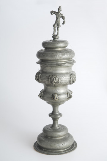 Puchar o okrągłej stopie, długim trzonie i wysokiej czarze nakrytej dwustopniową pokrywą, zwieńczoną figurką wojownika w antykizującej zbroi. Pokrywa lekko zdeformowana. Na wybrzuszeniach czaszy rzędy masek lwów do mocowania zawieszek.