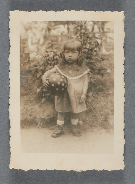 Fotografia monochromatyczna dziewczynki, około 2-3-letniej, z jasnymi włosami za szyję, w sukience, z ciemną piłką w jasne kropki pod pachą, stojącej obok krzewu. Ma wysokie buty i białe skarpetki. Stoi na wprost (ujęcie całopostaciowe), patrzy w obiektyw. Z opisu wynika, że jest to siostrzenica Mani Żelaznej, córka nieznanej nam z imienia siostry. Ta część rodziny została zabita w Zagładzie.Krawędzie ząbkowane.