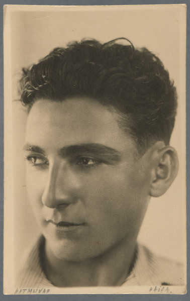 Fotografia portretowa młodego mężczyzny, półprofil, patrzy w lewo - część twarzy mniej widoczna mocno oświetlona, co nadaje zdjęciu charakter artystyczny; ciemne niedługie włosy postawione do góry. Widoczna prążkowana jasna koszula. Zdjęcie wywołane na papierze z nadrukiem pocztówkowym. Krawędzie odbitki proste, widoczny jest wzdłuż nich margines.