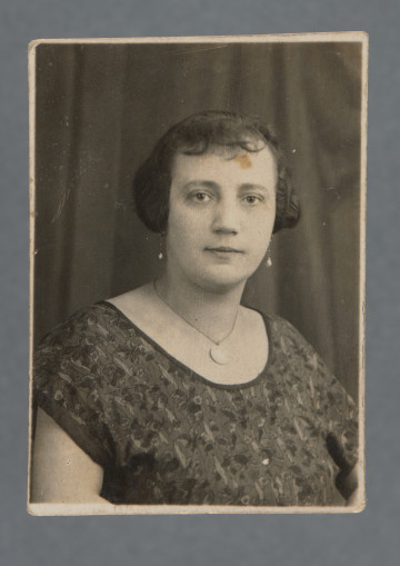Fotografia portretowa kobiety z medalionem na szyi, en face, na tle ciemnej pofałdowanej kotary. Ubrana w bluzkę z krótkimi rękawami, włosy za uszy, z grzywką.Krawędzie karty proste, widoczny cienki margines.
