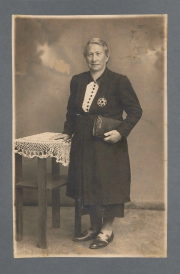 MPOLIN-A48.1.65 Fotografia monochromatyczna całopostaciowa Hany Hindy z d. Kleczewskiej, matki Hilela Poznera. W ciemnym kostiumie. Twarz uchwycona w trzech czwartych. Fotografia gabinetowa, karta z ząbkowaniem na krawędziach, na neutralnym tle, przy stoliku z obrusem, o który kobieta opiera się lewą ręką. W prawej trzyma torebkę.Na lewej piersi gwiazda Dawida - jako znak dyskryminacyjny. W czasie okupacji niemieckiej znalazła się w Wielkopolsce, w której przez wiele lat (do co najmniej 1918) żyła z mężem. Fotografia wykonana została w Koninie, dedykacja napisana w odległym o 30 km od Konina Zagórowie. Możemy przypuszczać, że do Zagórowa została przesiedlona z grupami innych Żydów z Konina, których część trafiła do getta w Zagórowie funkcjonującego w 1940–1942; przebywało w nim 2,5 tys. osób, rozstrzelanych później w pobliskich lasach kazimierzowskich - nie wiemy, czy kobieta zginęła w tym mordzie, czy trafiła do funkcjonującego na przełomie 1942/1943 obozu pracy przymusowej dla Żydów. Od darczyńcy wiadomo, że fotografia zachowała się, ponieważ została przesłana do rodziny w Portugalii - dedykacja z rewersu wskazuje, że wysyłana była do Salomona Perela i Fiszela.Na rewersie ślady po karcie albumu, do którego zdjęcie było przyklejone, przebarwienia po kleju.