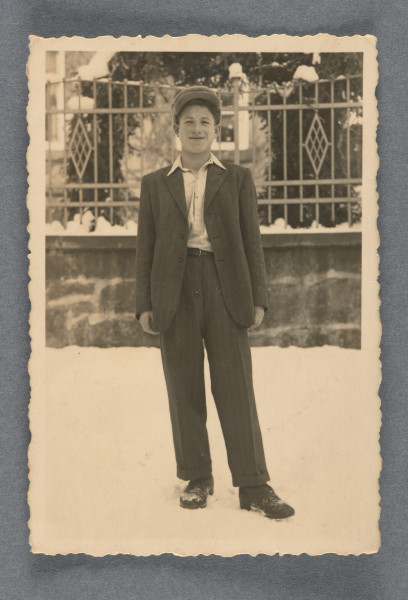 Fotografia nastoletniego chłopca w grubej czapce z daszkiem i samym garniturze i koszuli, stojącego na śniegu, ujętego na wprost, en face, uśmiechniętego. Stoi na tle ogrodzenia (płot na podmurówce) przed zabudowaniem. Na rewersie widoczny, pod dedykacją, numer - ołówkiem: 3610 Krawędzie odbitki ząbkowane, wzdłuż nich margines.