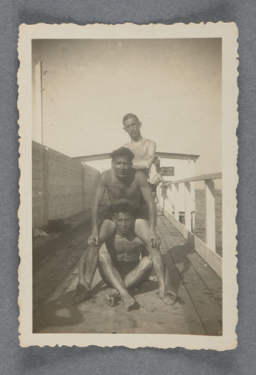 Fotografia monochromatyczna trzech młodych mężczyzn, jeden pochylony nad drugim, trzeci stojący za nimi - na drewnianym pomoście z balustradą, w tle widoczny biały budynek zapewne zamykający pomost.U dołu, siedzący z założonymi w goleniach nogami, Hilel Pozner. W tle widoczny oparty o balustradę inny mężczyzna, przypadkowo wszedł w kadr. U dołu po lewej cień fotografa(ki). Odbitka z prostokątnym marginesem białym. Krawędzie ząbkowane.