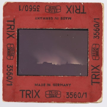 Zdjęcie płonącego getta warszawskiego: slajd w czerwonej ramce z czarnymi napisami - przy wszystkich krawędziach z każdej strony jednakowy: ,,MADE IN GERMANY | TRIX ZEISS | IKON [dwa ostatnie wyrazy obramowane cienką, miejscami zaokrąglaną linią; pośrodku ramki - pomiędzy nimi - także linia] 3560/1''; po jednej ze stron ramki przy dłuższej krawędzi uszkodzenie tektury - zdarty fragment o szerokości około 1 cm, papier w tym miejscu w kolorze zbliżonym do pomarańczowego. Wewnątrz ramki szybka, mocowana za pomocą blaszki.
Na przeźroczu, na obszernym czarnym tle - przy czym czerń wypełnia także boczne i dolną część kadru - widoczne żółtawe płomienie i wyżej, nieco od nich ciemniejsza, łuna. Kadr jest bardzo podobny do tego z j.a. MPOLIN-A2.2.7. Na górnym marginesie diapozytywu widoczny fragment napisu, po lewej: ,,[G]FACOLOR'' (napis firmowy - to błona agfacolor; pierwsza litera widoczna tylko częściowo), w prawym górnym rogu samo ,,A[G - litera widoczna tylko częściowo]''. Na dolnym marginesie: ,,25'' i ,,25 A''. Zdjęcie wykonane zostało nocą, z dachu wówczas siedmiopiętrowej (dziś czteropiętrowej) kamienicy narożnej przy pl. Dąbrowskiego u zbiegu Kredytowej i Jasnej (przy ambasadzie włoskiej). Na pierwszym planie, przed rozjaśnioną płomieniami przestrzenią, ciemne bryły budynków, których górna krawędź zaczyna się około 0,5 cm nad krawędzią obrazu. Po prawej, blisko środka szerokości, widoczna wieża, obok budynków z nierówną linią krawędzi górnej; przy drugiej krawędzi, lewej, obszerna budowla lub budowle o dachu raczej płaskim. Interpretacja: w centrum ujęcia znajduje się, jak można sądzić, sygnaturka kościoła św. Antoniego Padewskiego przy ul. Senatorskiej; po lewej widoczne byłyby gmachy: pałacu Komisji Rządowej Przychodów i Skarbu oraz pałacu Ministra Skarbu.
Na wcześniejszych etapach opracowywania fotografii wysuwano domniemanie, że właściwym awersem zdjęcia jest ta strona, na której wieża znajduje się po lewej stronie - a budowla po wówczas prawej stronie miała być gmachem Teatru Wielkiego. Jest to jednak interpretacja słabsza niż powyższa, zwłaszcza wobec napisów na diapozytywie wskazujących, jak się wydaje, jednoznacznie awers.