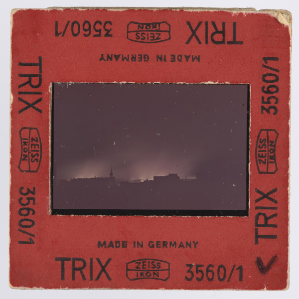 Zdjęcie płonącego getta warszawskiego: slajd w czerwonej ramce z czarnymi napisami - przy wszystkich krawędziach z każdej strony jednakowy: ,,MADE IN GERMANY | TRIX   ZEISS | IKON [dwa ostatnie wyrazy obramowane cienką, miejscami zaokrąglaną linią; pośrodku ramki - pomiędzy nimi - także linia]    3560/1''; w prawych dolnych rogach po obu stronach ramki - napisane na czarno: ,,V''. Wewnątrz ramki szybka, mocowana za pomocą blaszki.Na przeźroczu, na obszernym czarnym tle - przy czym czerń wypełnia także boczne i dolną część kadru - widoczne żółtawe płomienie i wyżej, nieco od nich ciemniejsza, łuna. Na górnym marginesie przeźrocza widoczny fragment napisu, po lewej: ,,FACOLOR'' (napis firmowy - to błona agfacolor), w prawym górnym rogu samo ,,A''. Na dolnym marginesie: ,,26'' i ,,26 A''. Zdjęcie wykonane zostało nocą, z dachu wówczas siedmiopiętrowej (dziś czteropiętrowej) kamienicy narożnej przy pl. Dąbrowskiego u zbiegu Kredytowej i Jasnej (przy ambasadzie włoskiej). Na pierwszym planie, przed rozjaśnioną płomieniami przestrzenią, ciemne bryły budynków, których górna krawędź zaczyna się około 0,5 cm nad krawędzią obrazu. Po prawej, blisko środka szerokości, widoczna wieża, obok budynków z nierówną linią krawędzi górnej; przy drugiej krawędzi, lewej, obszerna budowla lub budowle o dachu raczej płaskim. Interpretacja: w centrum ujęcia znajduje się, jak można sądzić, sygnaturka kościoła św. Antoniego Padewskiego przy ul. Senatorskiej; po lewej widoczne byłyby gmachy: pałacu Komisji Rządowej Przychodów i Skarbu oraz pałacu Ministra Skarbu.Na wcześniejszych etapach opracowywania fotografii wysuwano domniemanie, że właściwym awersem zdjęcia jest ta strona, na której wieża znajduje się po lewej stronie - a budowla po wówczas prawej stronie miała być gmachem Teatru Wielkiego. Jest to jednak interpretacja słabsza niż powyższa, zwłaszcza wobec napisów na diapozytywie wskazujących, jak się wydaje, jednoznacznie awers.