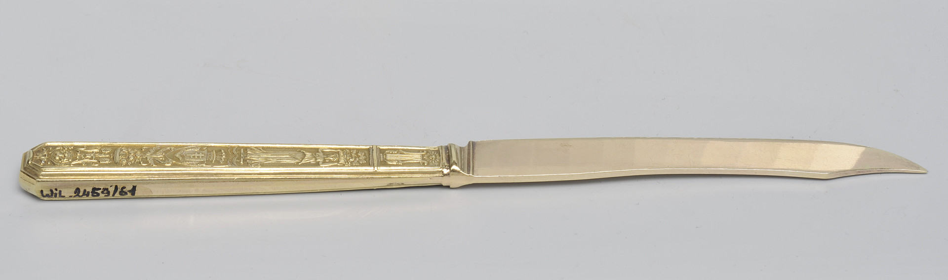 ujęcie 3/4 - strona z gładką głownią. Ujęcie z widocznym grzbietem. Czubek ostrza skierowany w prawą stronę. Na końcu górnej krawędzi trzonka numer inwentarzowy.