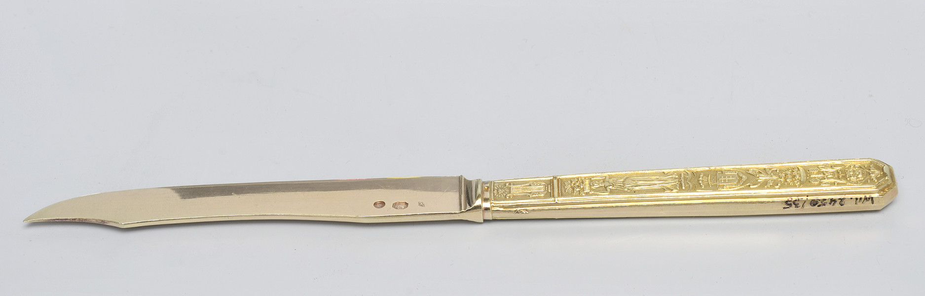 ujęcie 3/4 - strona z puncami na głowni. Trzy punce o różnych kształtach przy łączeniu głowni z rękojeścią. Ujęcie z widocznym grzbietem i szczytem ostrza skierowanym w lewo. Na końcu górnej krawędzi trzonka numer inwentarzowy.