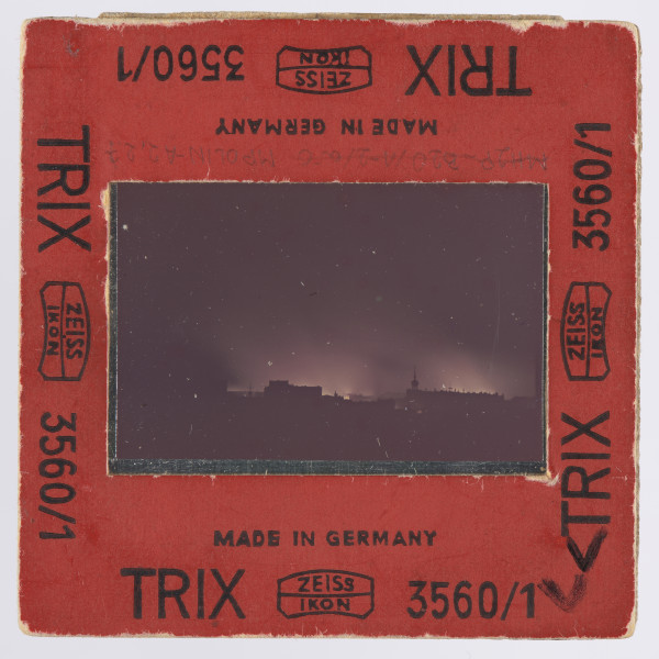 Zdjęcie płonącego getta warszawskiego: slajd w czerwonej ramce z czarnymi napisami - przy wszystkich krawędziach z każdej strony jednakowy: ,,MADE IN GERMANY | TRIX   ZEISS | IKON [dwa ostatnie wyrazy obramowane cienką, miejscami zaokrąglaną linią; pośrodku ramki - pomiędzy nimi - także linia]    3560/1''; w prawych dolnych rogach po obu stronach ramki - napisane na czarno: ,,V''. Wewnątrz ramki szybka, mocowana za pomocą blaszki.Na przeźroczu, na obszernym czarnym tle - przy czym czerń wypełnia także boczne i dolną część kadru - widoczne żółtawe płomienie i wyżej, nieco od nich ciemniejsza, łuna. Na górnym marginesie przeźrocza widoczny fragment napisu, po lewej: ,,FACOLOR'' (napis firmowy - to błona agfacolor), w prawym górnym rogu samo ,,A''. Na dolnym marginesie: ,,26'' i ,,26 A''. Zdjęcie wykonane zostało nocą, z dachu wówczas siedmiopiętrowej (dziś czteropiętrowej) kamienicy narożnej przy pl. Dąbrowskiego u zbiegu Kredytowej i Jasnej (przy ambasadzie włoskiej). Na pierwszym planie, przed rozjaśnioną płomieniami przestrzenią, ciemne bryły budynków, których górna krawędź zaczyna się około 0,5 cm nad krawędzią obrazu. Po prawej, blisko środka szerokości, widoczna wieża, obok budynków z nierówną linią krawędzi górnej; przy drugiej krawędzi, lewej, obszerna budowla lub budowle o dachu raczej płaskim. Interpretacja: w centrum ujęcia znajduje się, jak można sądzić, sygnaturka kościoła św. Antoniego Padewskiego przy ul. Senatorskiej; po lewej widoczne byłyby gmachy: pałacu Komisji Rządowej Przychodów i Skarbu oraz pałacu Ministra Skarbu.Na wcześniejszych etapach opracowywania fotografii wysuwano domniemanie, że właściwym awersem zdjęcia jest ta strona, na której wieża znajduje się po lewej stronie - a budowla po wówczas prawej stronie miała być gmachem Teatru Wielkiego. Jest to jednak interpretacja słabsza niż powyższa, zwłaszcza wobec napisów na diapozytywie wskazujących, jak się wydaje, jednoznacznie awers.