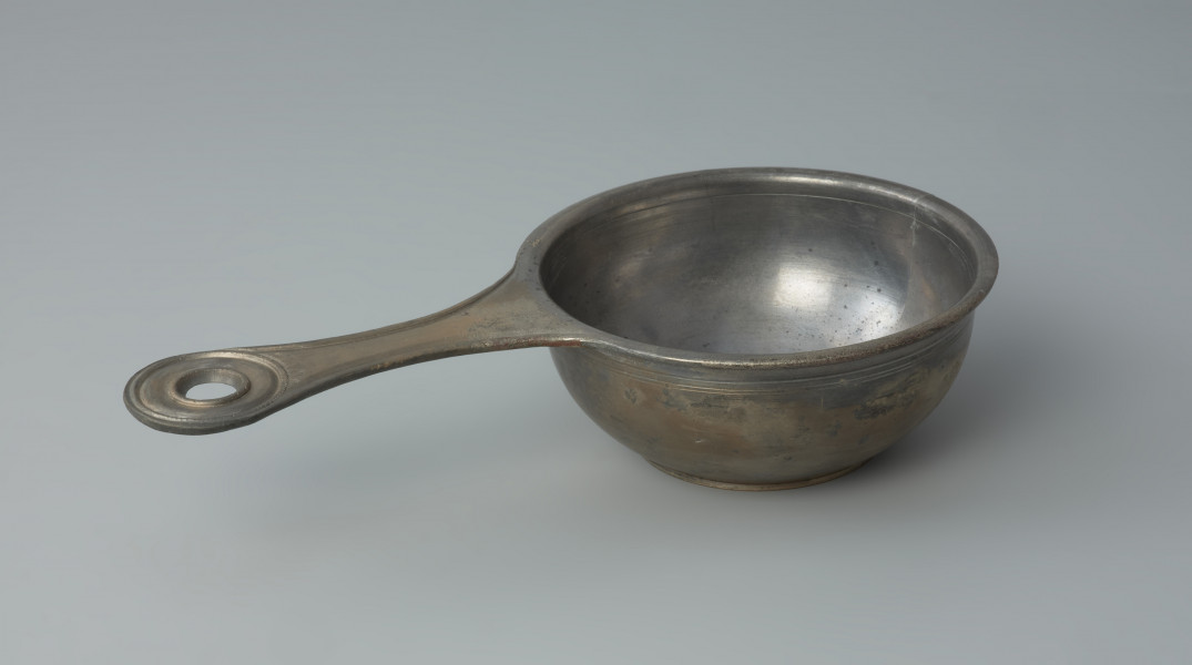 naczynie; Rzymski brązowy rondelek - Ujęcie z góry z prawego boku. Rzymski brązowy rondelek z płaskim dnem o przewężonym uchwycie z otworem. Rondel jest zdobiony pod wylewem płytkimi dookolnymi liniami rytymi.