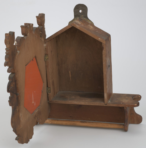 Pięciokątna szafka osadzona na prostokątnej podstawie. W dolnej części podstawy drążek. Drzwiczki do szafki z owalnym lustrem w rzeźbionej ramie o nieregularnym konturze, osadzone na dwóch zawiasach, zamykane na podłużny haczyk. W środku miejsce na schowanie drobnych rzeczy. Z dwóch stron lustra umieszczone są stojące lwy, z głowami odwróconymi na zewnątrz. Nad i pod lustrem rzeźbione motywy floralne i liście. Lwie grzywy oraz tło są w ciemniejszym odcieniu drewna. Na szczycie szafki, z tyłu, umieszczona została blaszana zawieszka. 