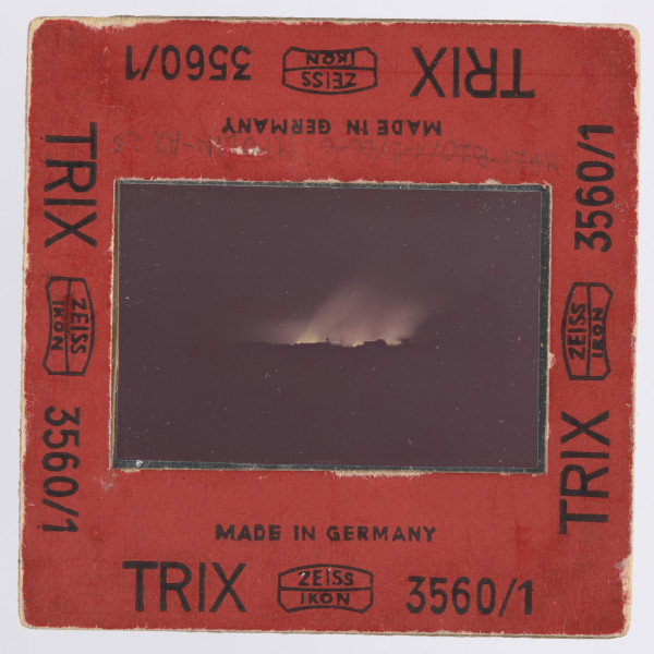 Zdjęcie płonącego getta warszawskiego: slajd w czerwonej ramce z czarnymi napisami - przy wszystkich krawędziach z każdej strony jednakowy: ,,MADE IN GERMANY | TRIX   ZEISS | IKON [dwa ostatnie wyrazy obramowane cienką, miejscami zaokrąglaną linią; pośrodku ramki - pomiędzy nimi - także linia]    3560/1''. Wewnątrz ramki szybka, mocowana za pomocą blaszki.Na przeźroczu, na obszernym czarnym tle - przy czym czerń wypełnia także około 1 cm dolnej części kadru i boki - widoczne żółtawe płomienie i wyżej, nieco od nich ciemniejsza, łuna, pochylona w lewo (na zachód). Na górnym marginesie przeźrocza, po lewej widoczny napis: ,,AGFACOLOR'' (napis firmowy). Na dolnym marginesie: ,,18'' i ,,18 A''. Zdjęcie wykonane zostało nocą, z dachu wówczas siedmiopiętrowej (dziś czteropiętrowej) kamienicy narożnej przy pl. Dąbrowskiego u zbiegu Kredytowej i Jasnej (przy ambasadzie włoskiej). Na pierwszym planie, przed rozjaśnioną płomieniami przestrzenią, ciemne bryły budynków, których górna krawędź zaczyna się około 1 cm nad krawędzią obrazu. Pośrodku widoczna wieża, obok budynków z nierówną linią krawędzi górnej; po lewej od niej obszerna budowla lub budowle o dachu raczej płaskim. Interpretacja: w centrum ujęcia znajduje się, jak można sądzić, sygnaturka kościoła św. Antoniego Padewskiego przy ul. Senatorskiej, a po lewej widoczne byłyby gmachy: pałacu Komisji Rządowej Przychodów i Skarbu oraz pałacu Ministra Skarbu.Na wcześniejszych etapach opracowywania fotografii wysuwano domniemanie, że właściwym awersem zdjęcia jest ta strona, na której wieża znajduje się po lewej stronie - a budowla po wówczas prawej stronie miała być gmachem Teatru Wielkiego. Jest to jednak interpretacja słabsza niż wyżej opisana, zwłaszcza jeśli wziąć pod uwagę napisy na diapozytywie, wskazujące, jak się wydaje, jednoznacznie awers.