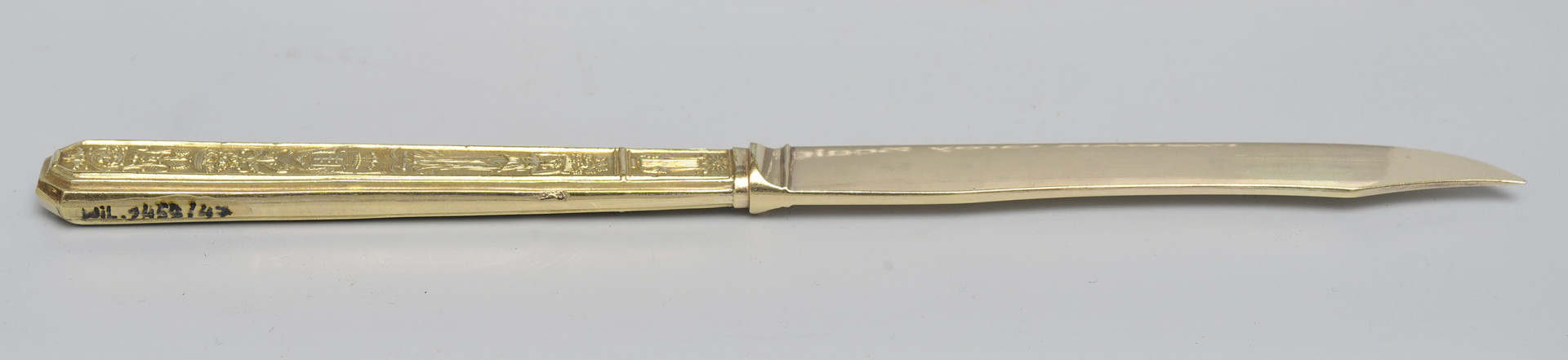 ujęcie 3/4 - strona z gładką głownią. Ujęcie z widocznym grzbietem. Czubek ostrza skierowany w prawą stronę. Na końcu górnej krawędzi trzonka numer inwentarzowy.