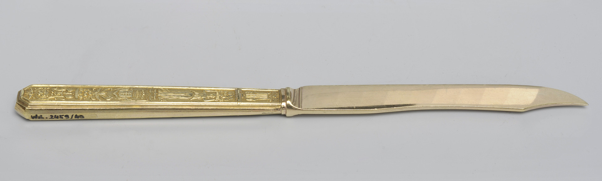 ujęcie 3/4 - strona z gładką głownią. Ujęcie z widocznym grzbietem i szczytem ostrza skierowanym w prawo. Na końcu górnej krawędzi trzonka numer inwentarzowy.