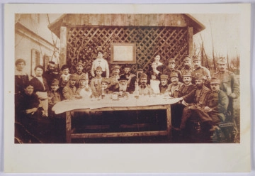MPOLIN-A13.2.1 Fotografia rodzinna z austriackimi żołnierzami - Zdjęcie grupowe wykonane w czasie święta Pesach. Wtórna odbitka. Ozjasz Grossbart (siedzi pośrodku stołu), dziadek Emilii Leibel, zaprosił żołnierzy austriackich pochodzenia żydowskiego na wspólne obchody. Jeden z żołnierzy ma na rękawie opaskę z czerwonym krzyżem. Na zdjęciu widoczne także kobiety: jedna z nich, w fartuchu, trzyma talerz, jakby zatrzymała się na moment zrobienia zdjęcia w trakcie nakrywania do stołu, inna kobieta, w ciemnej sukni, siedząca, trzyma chochlę. W tle widoczny obraz, o którym wiemy z relacji, że był podobizną cesarza Franciszka Józefa. Uroczystość zorganizowano na zewnątrz, stół ustawiono na tle drewnianej ściany konstrukcji w formie pergoli.