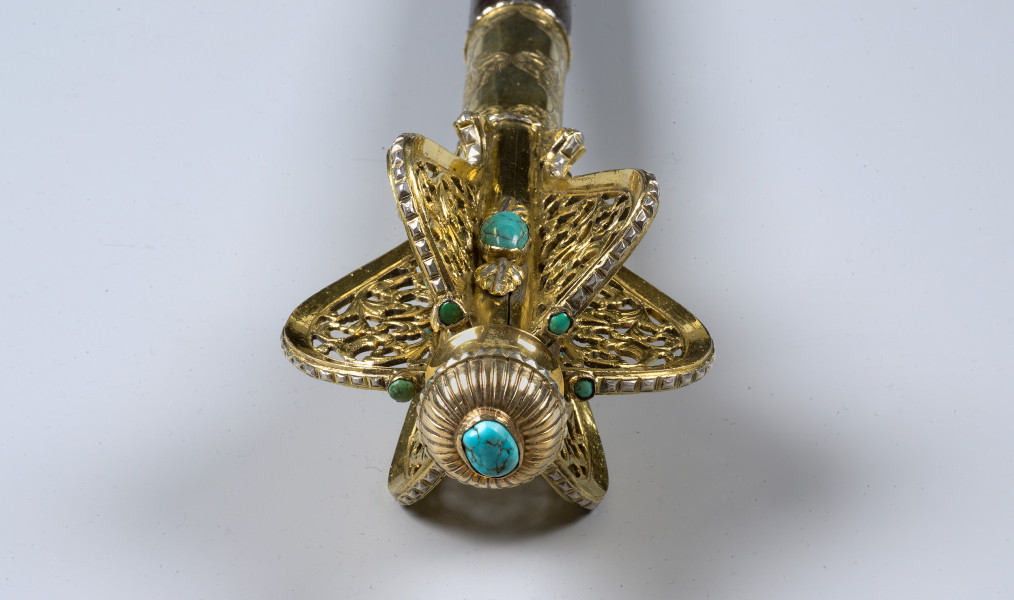 zbliżenie na głowicę złożoną z 6 ażurowych piór, między którymi osadzone turkusy - ujęcie z góry. Złocone pióra promieniście rozchodzące się od rękojeści.