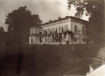 Fotografia prezentuje pałac wraz z otaczającą go bujną roślinnością od strony południowo-zachodniej. Wokół  pałacu widoczne są nowe nasadzenia drzew liściastych. 