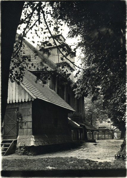 Na zdjęciu znajduje się drewniany kościół między drzewami.