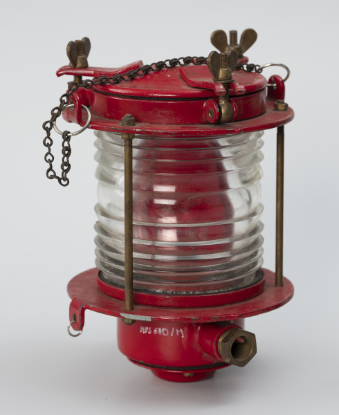 Elektryczna latarnia okrętowa - ujęcie z drugiej strony; Latarnia elektryczna od góry i dołu wyposażona w dwie cylindryczne, metalowe części, osadzone na okrągłych tarczach, które połączone są trzema prętami o średnicy 4 mm. Cylindryczna część środkowa z białego szkła o średnicy 100 mm i wysokości 110 mm stanowi soczewkę Frensela. Dolna część lampy o średnicy 80 mm i wysokości 60 mm z dławikiem przedzielona jest na dwie części uszczelnione gumową uszczelką. Spodnie wieko przykręcone na trzy śruby. Część górna o średnicy 150 mm i wysokości 30 mm, z wiekiem oddzielonym uszczelką mocowanym na 3 śruby dociskowe na zawiasach z pokrętłami. Wewnątrz lampy czerwony filtr oraz żarówka mocowana do oprawki bagnetowej (B22d). Na obu metalowych częściach po 2 uchwyty, umieszczone na przeciwko siebie. W górnej części przymocowano do nich metalowe kółka połączone łańcuchem.