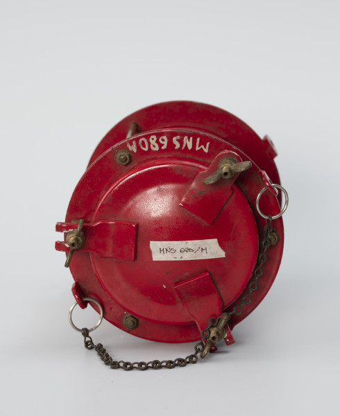 Elektryczna latarnia okrętowa - ujęcie z góry; Latarnia elektryczna. Część górna o średnicy 150 mm i wysokości 30 mm, z wiekiem oddzielonym uszczelką mocowanym na 3 śruby dociskowe na zawiasach z pokrętłami. Na widoczne 2 uchwyty, umieszczone na przeciwko siebie. W górnej części przymocowano do nich metalowe kółka połączone łańcuchem.