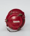 Elektryczna latarnia okrętowa - ujęcie z góry; Latarnia elektryczna. Część górna o średnicy 150 mm i wysokości 30 mm, z wiekiem oddzielonym uszczelką mocowanym na 3 śruby dociskowe na zawiasach z pokrętłami. Na widoczne 2 uchwyty, umieszczone na przeciwko siebie. W górnej części przymocowano do nich metalowe kółka połączone łańcuchem.