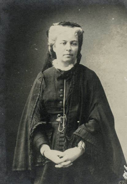 Na zdjęciu znajduje się kobieta w ciemnej sukni; jasne włosyupięte do tyłu.