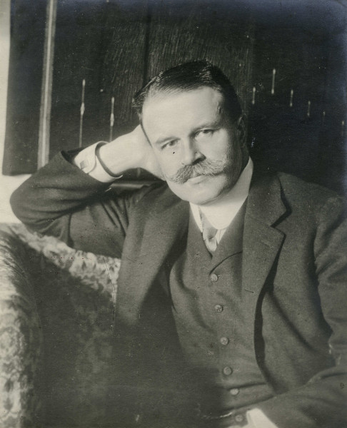 Na zdjęciu znajduje się mężczyzna z wąsami, zaczesanymi na prawy jego bok włosami, ubrany w garnitur, białą koszulę i krawat, podpiera ręką głowę.