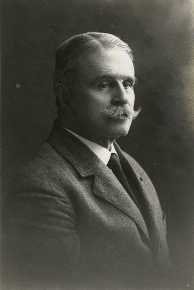 Na zdjęciu widoczny jest mężczyzna w marynarce i krawacie. Włosy, zaczesane ma na boki i ma gęste wąsy