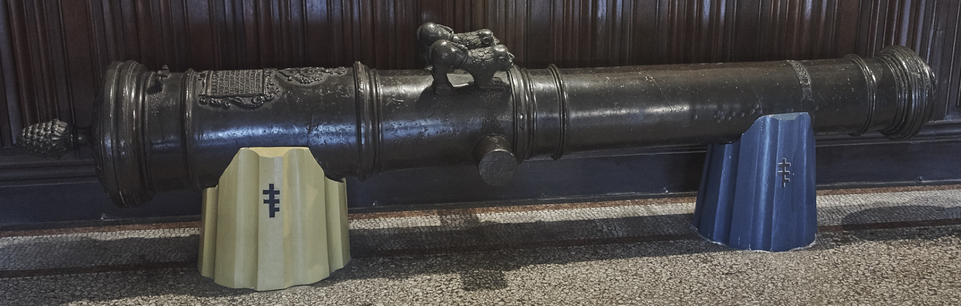 Na zdjęciu znajduje się lufa armatnia wsparta na dwóch drewnianych postumentach ozdobionych herbem Pilawa.