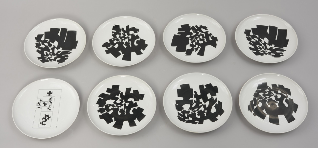 talerze - Ujęcie wszystkich ośmiu talerzy z góry. Zestaw ośmiu białych talerzy, na każdym nadrukowane czarne, geometryczne, abstrakcyjne formy, przypominające krzyże o zróżnicowanych proporcjach. Powstałe z przekształceń fotografii miasta (ulic, budynków) zajmują w różnych konfiguracjach różną część powierzchni każdego z ośmiu talerzy. Na jednym nadruk jest wyraźnie inny. W prostokącie o cienkim konturze znajdują się kolejne cienko konturowe prostokąty, trzy większe w jednym rzędzie oraz trzy mniejsze w drugim rzędzie. Te z kolei zawierają układy geometrycznych form, nawiązujące do uabstrakcyjnionych krzyży na pozostałych talerzach. W jednym z prostokątów krzyż równoramienny.