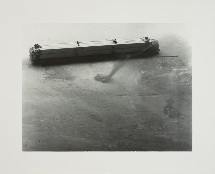 fotografia; W pracowni Apellesa XIII - Ujęcie z przodu na białym tle. Czarno-biała, pozioma kompozycja rozświetlona z góry, ciemniejsza u dołu. Na porysowanej i poplamionej podłodze z płytek pcv leży podłużny obiekt, ujęty z góry pod kątem. Jego lewy koniec przesunięty jest nieznacznie w kierunku pierwszego planu. Obiekt składa się z dwóch wiązek drewnianych listew, ułożonych jedna na drugiej i z płótna, połączonych za pomocą sznurka oraz śrub. W prawej części kadru ledwo widoczny, rozmazany w wyniku długiego naświetlania kliszy, zarys skulonej postaci, opierającej się szeroko rozstawionymi rękoma o podłogę.