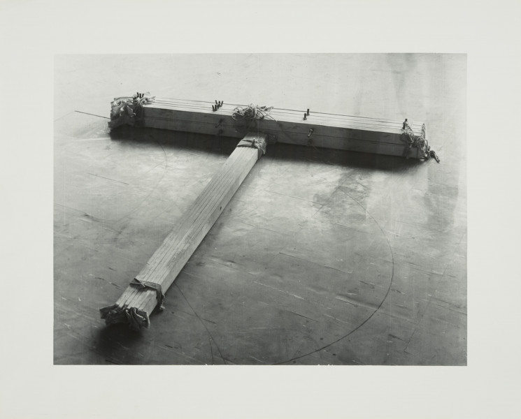 fotografia; W pracowni Apellesa XI - Ujęcie z przodu wraz z tłem. Czarno-biała, pozioma kompozycja rozświetlona od góry, ciemniejąca u dołu. Na podłodze z płytek pcv leży obiekt w kształcie litery T, ujęty z góry, pod kątem, z częścią horyzontalną lekko przekręconą ku prawej dolnej części kadru. Obiekt składa się z dwóch wiązek drewnianych listew i płótna, połączonych za pomocą sznurka oraz śrub. Na końcu wiązki listew ułożonych wertykalnie, schodzących do lewego dolnego rogu pracy, wystają wyraźnie kawałki płótna. Podłoga ciemnoszara, poprzecierana, porysowana gdzieniegdzie czarnymi nieregularnymi liniami.