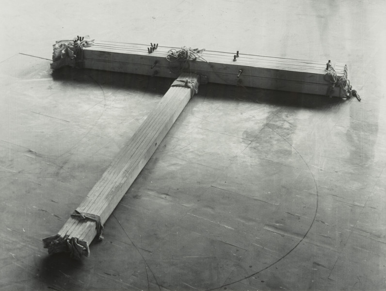 fotografia; W pracowni Apellesa XI - Ujecie z przodu bez tła. Czarno-biała, pozioma kompozycja rozświetlona od góry, ciemniejąca u dołu. Na podłodze z płytek pcv leży obiekt w kształcie litery T, ujęty z góry, pod kątem, z częścią horyzontalną lekko przekręconą ku prawej dolnej części kadru. Obiekt składa się z dwóch wiązek drewnianych listew i płótna, połączonych za pomocą sznurka oraz śrub. Na końcu wiązki listew ułożonych wertykalnie, schodzących do lewego dolnego rogu pracy, wystają wyraźnie kawałki płótna. Podłoga ciemnoszara, poprzecierana, porysowana gdzieniegdzie czarnymi neregularnymi liniami.