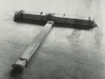 fotografia; W pracowni Apellesa XI - Ujecie z przodu bez tła. Czarno-biała, pozioma kompozycja rozświetlona od góry, ciemniejąca u dołu. Na podłodze z płytek pcv leży obiekt w kształcie litery T, ujęty z góry, pod kątem, z częścią horyzontalną lekko przekręconą ku prawej dolnej części kadru. Obiekt składa się z dwóch wiązek drewnianych listew i płótna, połączonych za pomocą sznurka oraz śrub. Na końcu wiązki listew ułożonych wertykalnie, schodzących do lewego dolnego rogu pracy, wystają wyraźnie kawałki płótna. Podłoga ciemnoszara, poprzecierana, porysowana gdzieniegdzie czarnymi neregularnymi liniami.