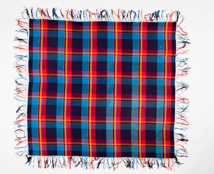 E/126/MRK/ML - Chustka kratówka wełniana. W kolorach: niebieski, czerwony, brązowy i czarny. Na brzegach kolorowe frędzle. Długość frędzli 14 cm. Wymiary chusty 160 x 150 cm. Wymiary krat: większe 12,5 x 11,5, najmiejsza 2 x 1,5 cm. Chusta częściowo podziurawiona. 