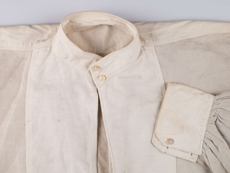 E/128/MRK/ML - Koszula lniana męska, biała, prosty i krótki kołnierzyk zapinany na 2 guziki, rękawy długie zapinane na 2 guziki. Dł. 101 cm, szer. 54 cm. Dł. rękawa 57 cm, szer. 20 cm, dł. mankietu 5 cm, szer. 11 cm. Z przodu koszuli rozcięcie  dł. 37 cm.  