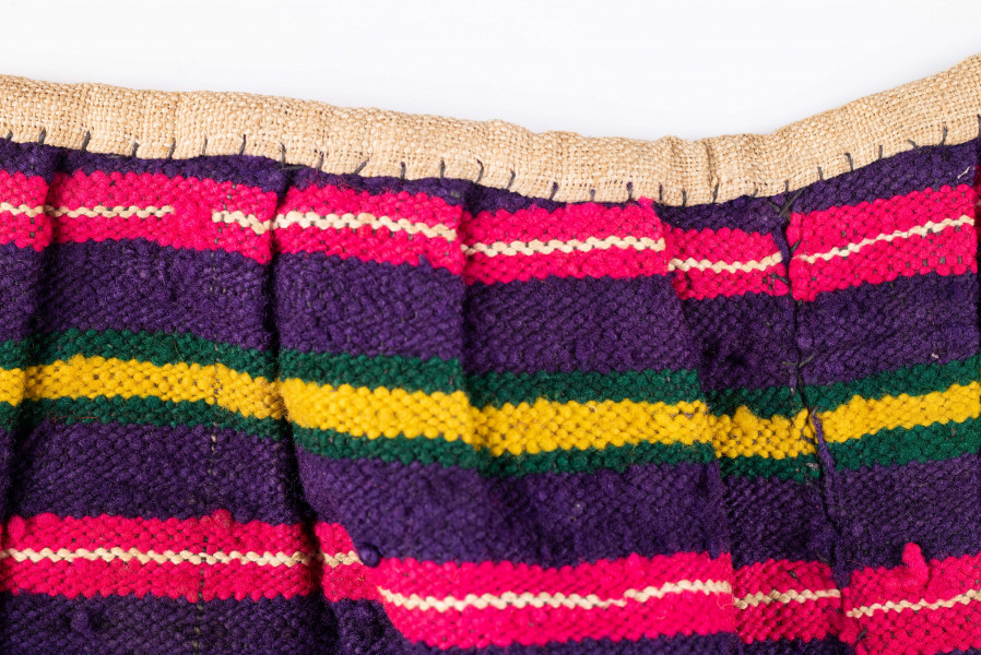 E/125/MRK/ML - Zapaska wełniana w różnokolorowe pasy, ułożone poziomo. Dł. zapaski 77 cm, szer. 98 cm, zszyta w środku. Kolory: fioletowy, niebieski, czerwony, biały, zielony, żółty, brązowy. Od góry 14 zakładek na nich naszyty lniany pasek szer. 1 cm, dł. 65 cm.