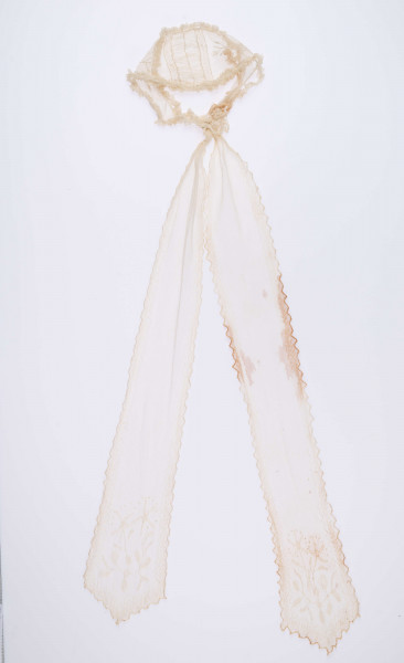 E/124/MRK/ML - Czepek tiulowy, biały tiul, wzory o motywach roślinnych, brzegi pofałdowane. Dług. wstążek 45 cm, szer. 8 cm, brzegi obszyte białą koronką szer. 1 cm. Czepek dł. 50 cm szer. 18 cm. W części środkowej wstawka szer. 10 cm.