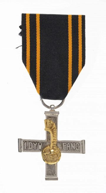 Odznaką Krzyża 1 Dywizji Pancernej jest krzyż bity w białym metalu o wymiarach 4,1 x 4,0 cm i szerokości ramion 0,7 cm. Powierzchnia krzyża z obydwu stron chropowata, obramowana wokół gładką ramką  szer. 0,1 cm. Na poziomych ramionach wypukły napis: 1 DYW. PANC. Na ramionach pionowych nałożona złocona miniaturka odznaki rozpoznawczej dywizji - szyszak na tle okrągłej tarczy ze skrzydłem husarskim. Krzyż noszony jest na czarnej wstążce szerokości 3,8 cm z 4 pomarańczowymi prążkami biegnącymi przy jej krawędziach.