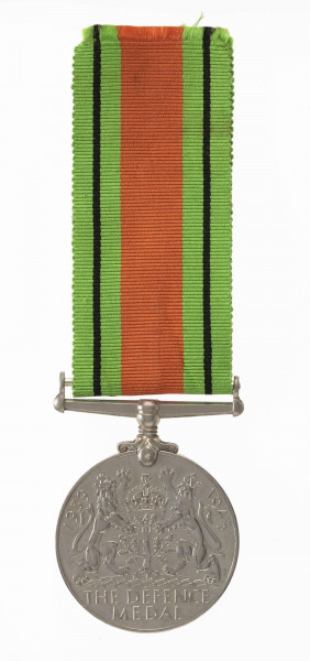 H/2050/MRK/ML - Odznaką jest medal bity w miedzioniklu 3,6 cm średnicy, o gładkim rancie. Na awersie jest lewy profil głowy króla Jerzego VI, przy zarycie szyi sygnatura HP. W otoku napis: GREGORIUS VI D:G:BR:OMN:REX F:D:IND:IMP. Na rewersie dąb zwięczony koroną zamkniętą, oraz dwa ryczące, wspięte na tylnych łapach lwy, za ich grzbietami daty 1939 i 1945. Dąb wyrasta z lądu oblanego wodą (wyspa). Poniżej w dwóch wierszach napis: THE DEFENCE MEDAL. Przywieszka w formie szlufy na wstążkę nitowana z medalem. Wstążka szerokości 3,2 cm, od brzegów jasnozielone pasy z czarnym prążkiem, w środku pas pomarańczowo-ceglasty.