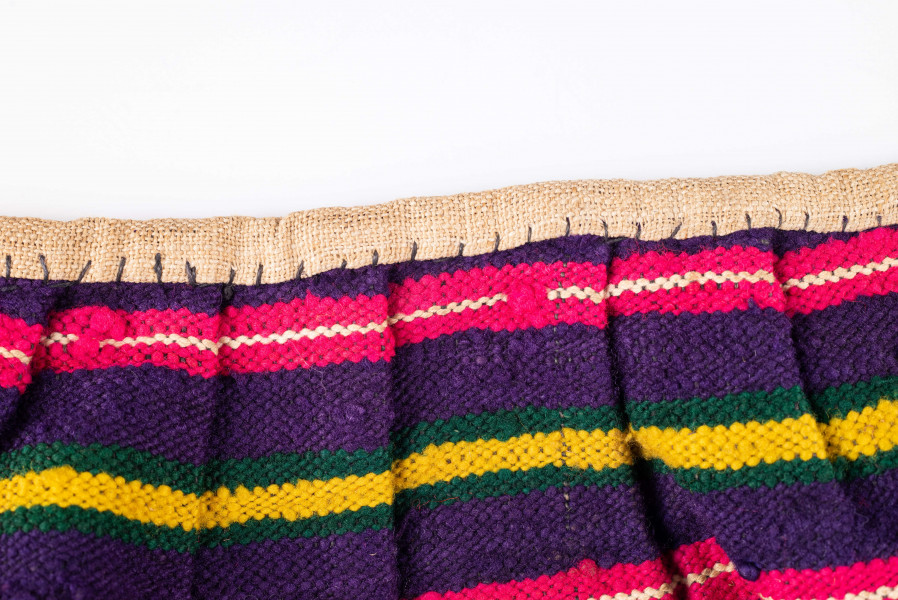 E/125/MRK/ML - Zapaska wełniana w różnokolorowe pasy, ułożone poziomo. Dł. zapaski 77 cm, szer. 98 cm, zszyta w środku. Kolory: fioletowy, niebieski, czerwony, biały, zielony, żółty, brązowy. Od góry 14 zakładek na nich naszyty lniany pasek szer. 1 cm, dł. 65 cm.