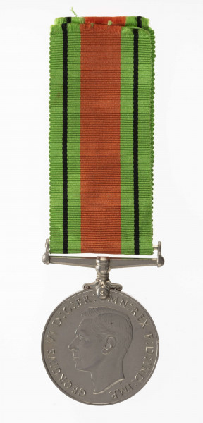 H/2050/MRK/ML - Odznaką jest medal bity w miedzioniklu 3,6 cm średnicy, o gładkim rancie. Na awersie jest lewy profil głowy króla Jerzego VI, przy zarycie szyi sygnatura HP. W otoku napis: GREGORIUS VI D:G:BR:OMN:REX F:D:IND:IMP. Na rewersie dąb zwięczony koroną zamkniętą, oraz dwa ryczące, wspięte na tylnych łapach lwy, za ich grzbietami daty 1939 i 1945. Dąb wyrasta z lądu oblanego wodą (wyspa). Poniżej w dwóch wierszach napis: THE DEFENCE MEDAL. Przywieszka w formie szlufy na wstążkę nitowana z medalem. Wstążka szerokości 3,2 cm, od brzegów jasnozielone pasy z czarnym prążkiem, w środku pas pomarańczowo-ceglasty.