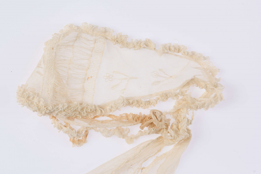 E/124/MRK/ML - Czepek tiulowy, biały tiul, wzory o motywach roślinnych, brzegi pofałdowane. Dług. wstążek 45 cm, szer. 8 cm, brzegi obszyte białą koronką szer. 1 cm. Czepek dł. 50 cm szer. 18 cm. W części środkowej wstawka szer. 10 cm.