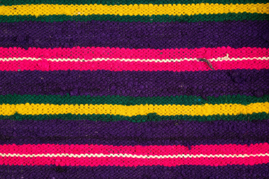 E/125/MRK/ML - Zapaska wełniana w różnokolorowe pasy, ułożone poziomo. Dł. zapaski 77 cm, szer. 98 cm, zszyta w środku. Kolory: fioletowy, niebieski, czerwony, biały, zielony, żółty, brązowy. Od góry 14 zakładek na nich naszyty lniany pasek szer. 1 cm, dł. 65 cm.