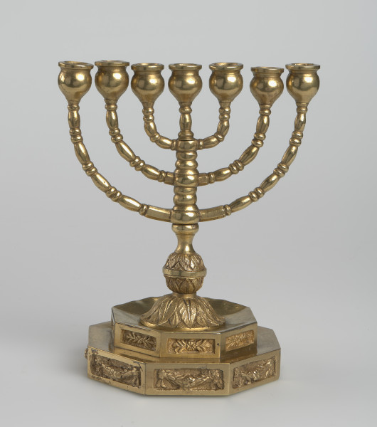 Świecznik - Podstawa ośmioboczna, dwustopniowa, o krawędzi dekorowanej panelami z reliefowym motywem roślinnym. Trzon tralkowy, zwieńczony wazonikową tulejką. Z trzonka symetrycznie wyprowadzone trzy pary łukowatych, tralkowych ramion zakończonych wazonikowymi tulejkami. Wszystkie tulejki ułożone w linii, na jednej wysokości.