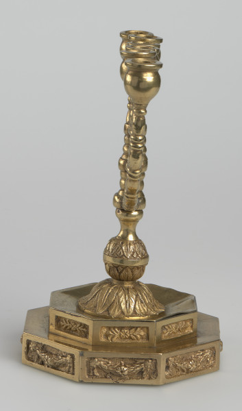 Świecznik - Podstawa ośmioboczna, dwustopniowa, o krawędzi dekorowanej panelami z reliefowym motywem roślinnym. Trzon tralkowy, zwieńczony wazonikową tulejką. Z trzonka symetrycznie wyprowadzone trzy pary łukowatych, tralkowych ramion zakończonych wazonikowymi tulejkami. Wszystkie tulejki ułożone w linii, na jednej wysokości.